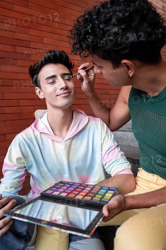 A joyful gay couple sharing a tender moment as one applies makeup to the other's face. They sit together against a brick wall, embracing diversity and self-expression
