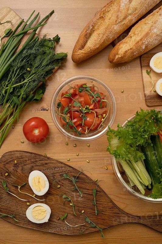 A top-down view of fresh ingredients for a salad, including herbs, tomato salad, boiled eggs, lettuce, baguettes, and green onions, beautifully arranged on a wooden surface