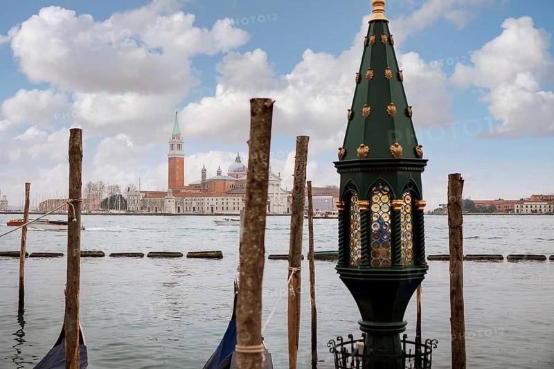 A tranquil view of Venice featuring a scenic waterfront with ornate architecture, wooden poles, and calm waters under a vibrant sky. Iconic landmarks subtly enhance the background