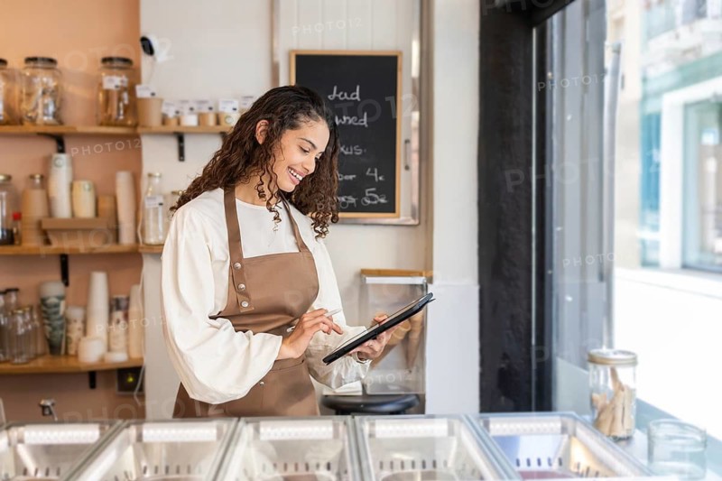 A cheerful woman clerk wearing a brown apron uses a tablet to manage orders in a modern ice cream shop. The bright interior and natural light enhance the pleasant atmosphere
