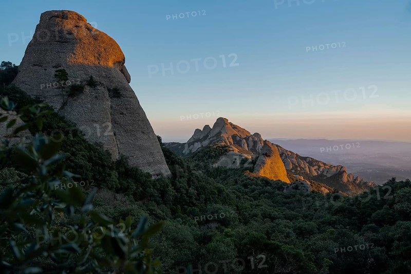 Dramatic rocky formations bathed in the warm glow of the setting sun, casting shadows over lush green vegetation in Montserrat, Catalonia, Spain. A serene natural beauty
