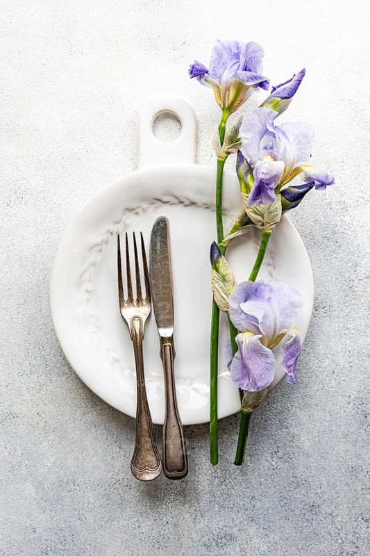 Top view of a sophisticated table setting featuring a white ceramic plate, vintage silverware, and vibrant iris flowers, arranged elegantly on a speckled grey background