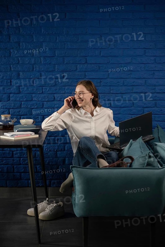 A woman in a white shirt and jeans sits on a couch, multitasking with a laptop and phone, surrounded by books and tea. The blue brick wall adds a vibrant backdrop