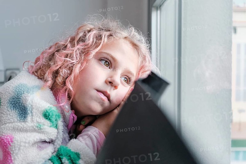 A young child with curly pink hair looks thoughtfully out a window. Wrapped in a cozy, colorful sweater, they display a serene and introspective expression in natural light at home