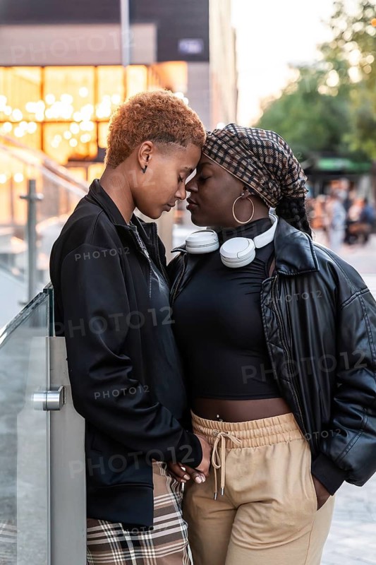 A tender moment between an LGBT multiethnic couple standing closely in an urban setting. They lovingly lean into each other, exuding warmth and connection amidst the city backdrop