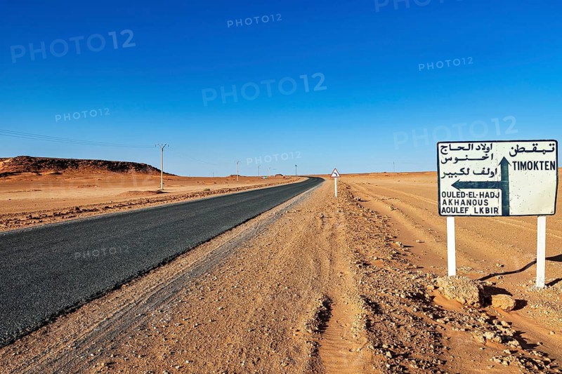 Road leads through a barren desert landscape under a clear blue sky with a signpost in Arabic, By car through the Sahara desert in Algeria