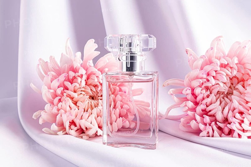 A square glass perfume bottle elegantly placed between two vibrant pink chrysanthemums on a delicate white silk fabric. The composition exudes luxury and femininity