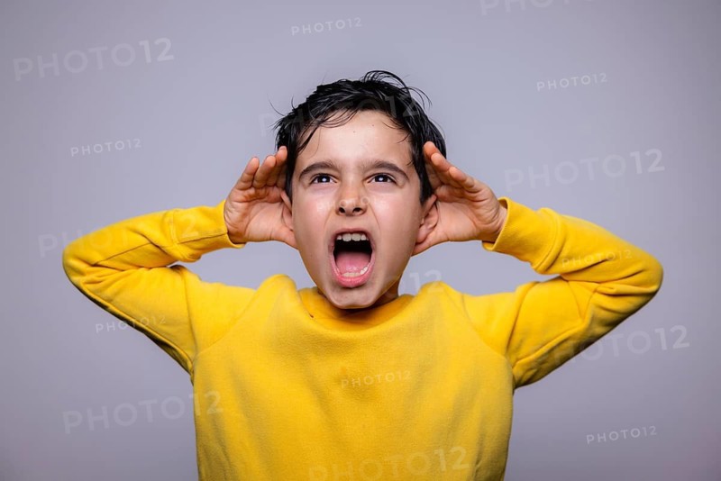 A young boy in a yellow sweater energetically shouts with hands raised to his ears, expressing a vibrant emotion against a plain background. The image captures youthful energy