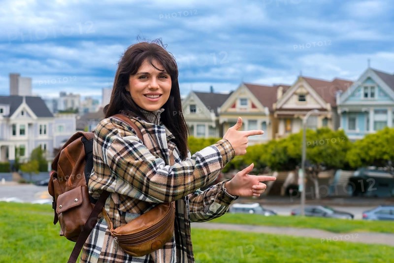 Young woman tourist visiting alamo square, pointing to the iconic victorian houses with a cityscape in the background, representing travel and urban exploration