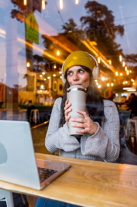 Young woman wearing headphones and yellow beanie working on her laptop in a san francisco cafe, enjoying a drink while looking out the window, reflecting the city lights