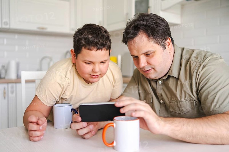 A father and son sit at a kitchen table, both focused on a smartphone. They share quality time, sipping from mugs and enjoying a moment of digital entertainment