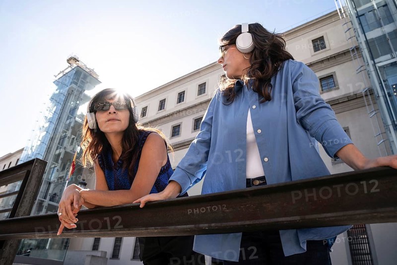 Multiethnic lesbian couple listens to music with headphones while enjoying the sunshine in a modern cityscape. Dressed stylishly, they embrace the moment with a mix of relaxation and curiosity