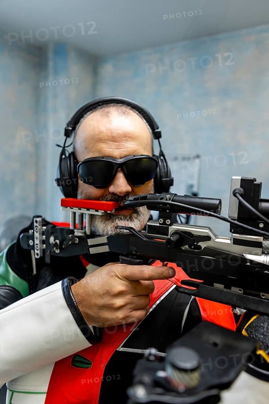 Blind athlete wearing headphones and specialized gear aims a rifle during an Olympic shooting event, highlighting skill, focus, and adaptive sports technology