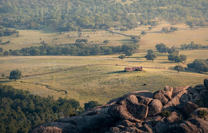 A breathtaking view from rocky ledge overlooking a verdant valley in the Guadarrama mountain range. Scattered trees and an abandoned house punctuate the expansive landscape in the Pedriza
