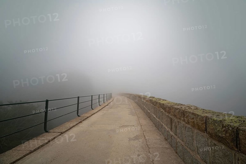 A serene stone bridge extends into thick fog, creating a mysterious and tranquil atmosphere. The fog obscures the horizon, inviting viewers to imagine what lies beyond