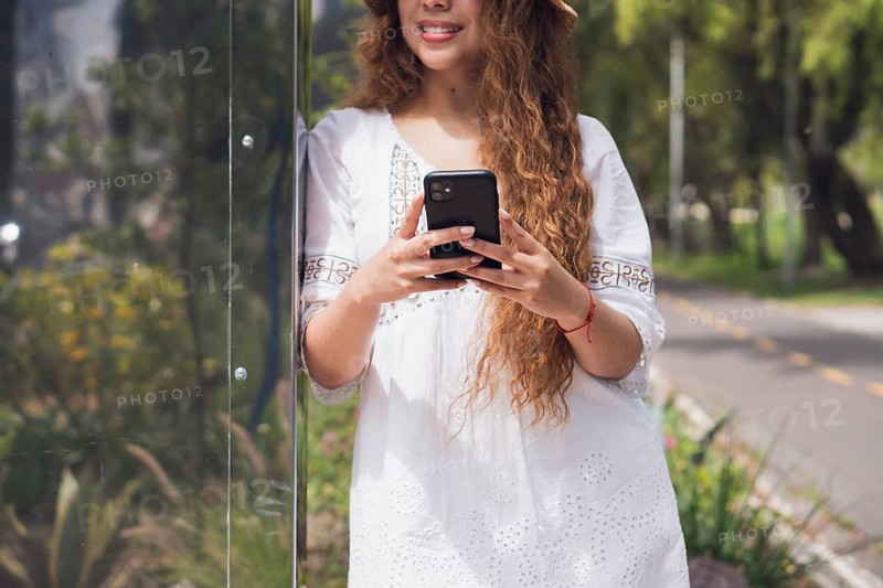 A young Latin American woman in a casual white dress stands outdoors using her smartphone. She leans against a reflective surface, enjoying a sunny day in the park