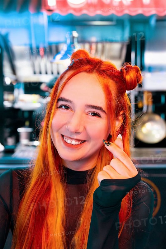 A smiling woman with vibrant red-orange hair at an Asian food market She gestures enthusiastically amidst market lights, capturing the lively and cultural environment