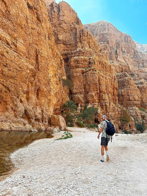 A male hiker walks along the rocky terrain of Wadi Ash Shab, a picturesque oasis nestled in Oman, showcasing the stunning natural landscapes and inviting exploration