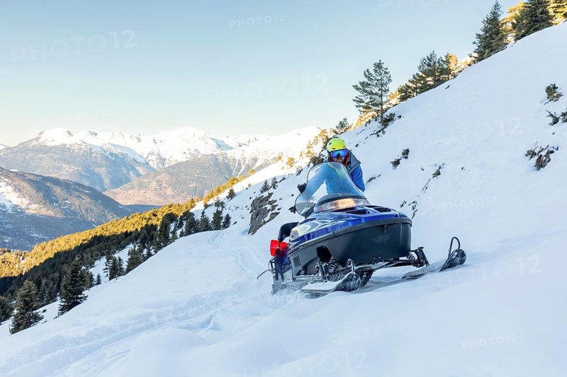 A man rides a snowmobile across a snowy mountain landscape, enjoying a winter adventure. The sun shines brightly over the scenic backdrop of evergreen trees and distant peaks