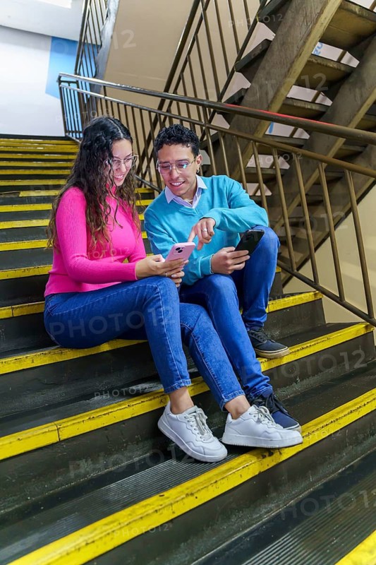 A young woman and a young man, both Gen Z, sit on a staircase sharing a moment with their smartphones. They are engaged in a modern, digital lifestyle, enjoying social interactions