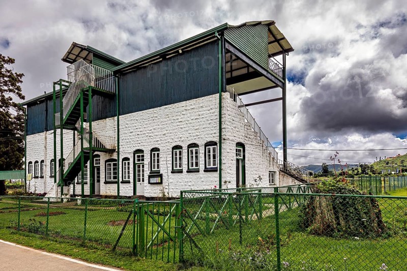 Historic building with outdoor stairs and sports stand surrounded by green areas and cloudy skies, The Nuwara Eliya Sri Lanka Racecourse
