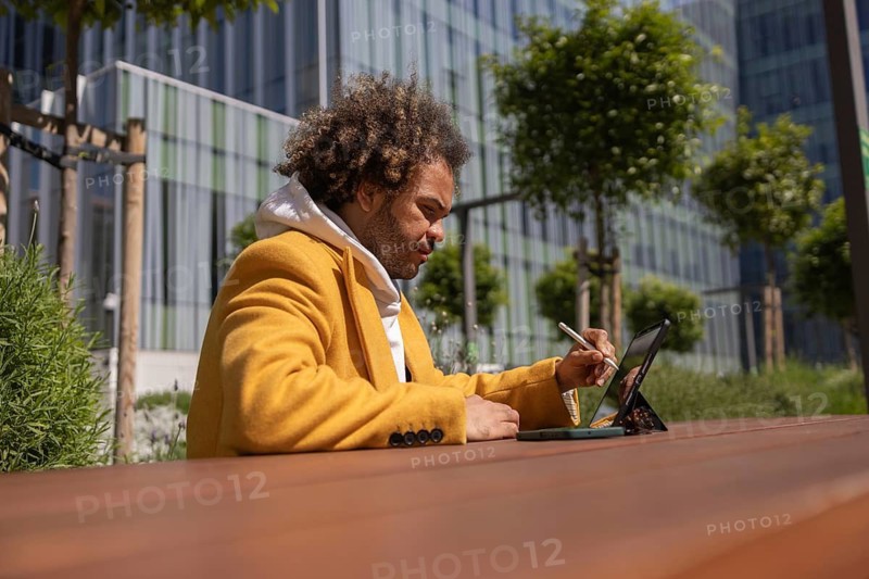 A man with afro hair in a yellow coat uses a tablet outdoors. Seated at a wooden table, he appears focused and relaxed, surrounded by greenery and modern architecture