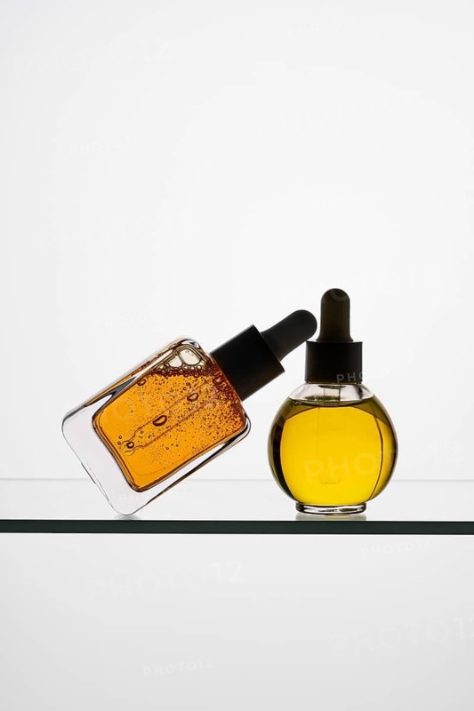Two pipette bottles filled with vitamin C serum rest on a glass shelf One bottle leans against the other, highlighting their golden hues against a minimalist white background