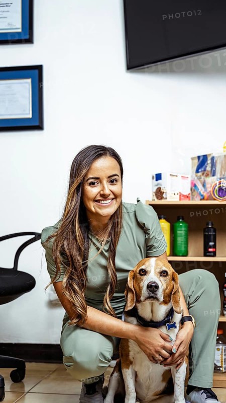 A nutritionist in a green uniform poses with her dog in a professional office setting. Certificates and health products are visible, emphasizing a focus on wellness and nutrition