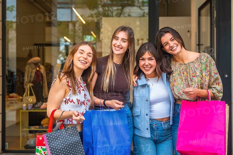 Four smiling young women friends posing in front of a clothing store, holding colorful shopping bags and enjoying their retail therapy experience together