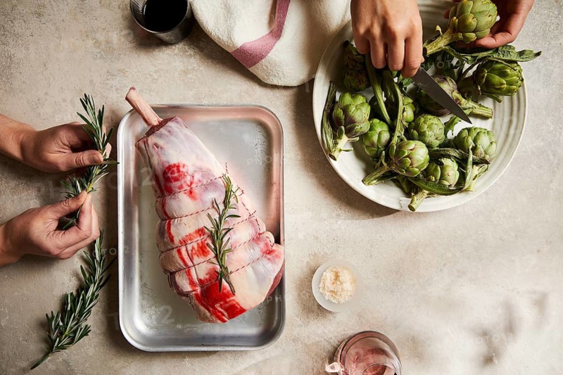 Butchers prepare a raw leg of lamb on a tray with fresh rosemary. Nearby, artichokes are pruned, highlighting the dedication and culinary skill in food preparation