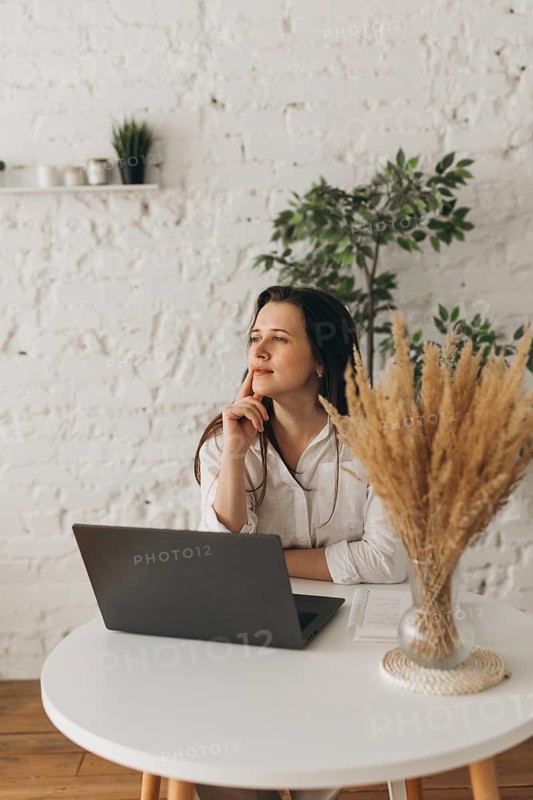 A nutritionist sits thoughtfully at a white desk with a laptop and pampas grass in a vase. The bright space is complemented by a brick wall and green plant decor