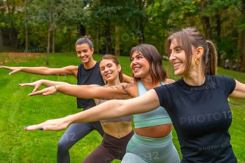 Group of smiling women practicing yoga together in a green park, doing warrior poses and stretches outdoors, enjoying fitness, friendship, and summer wellbeing