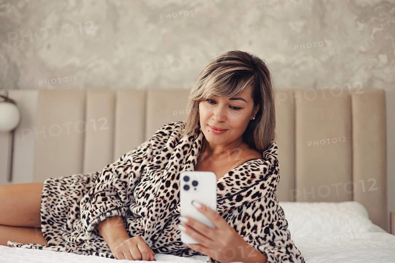 A woman in a leopard print robe lies comfortably on a bed while using her smartphone, capturing a moment of relaxation and technology in a cozy bedroom setting