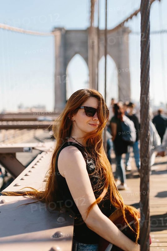 Young woman with long hair and sunglasses relaxing on a bridge during a sunny day in New York. Urban scenery with people in the background. Perfect for travel and lifestyle themes