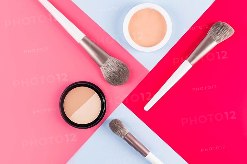 Top view of makeup brushes and face powders arranged on a vibrant red, pink and blue geometric background. Ideal for beauty, fashion, and cosmetic themes
