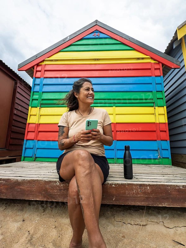 A woman with a smartphone sits relaxed in front of vibrant Brighton Bathing Boxes in Melbourne, Australia. The iconic, colorful beach huts create a lively backdrop