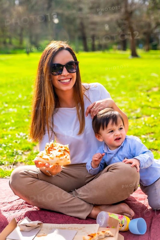 Happy mother sharing a slice of pizza with her son during a sunny picnic in the park, enjoying quality time and laughter together