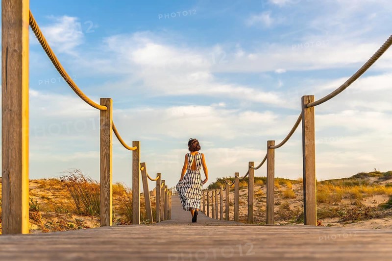 Woman walking away on a wooden boardwalk through sand dunes toward the atlantic beach at seignosse under a sunny summer sky, evoking freedom, holiday escape and calm horizons