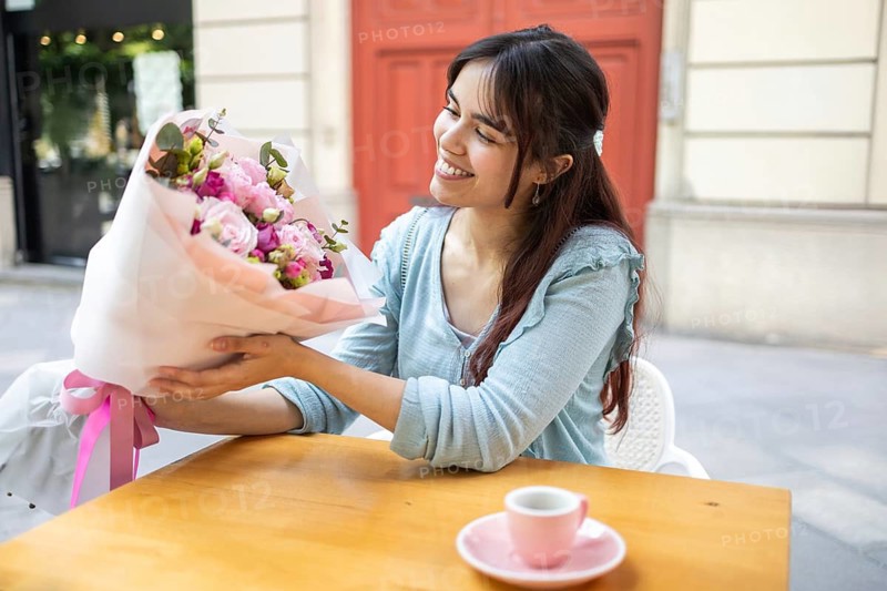 A smiling woman admires a vibrant bouquet of flowers wrapped in pink paper, which she bought at a local florist, while sitting at a cafe table with a coffee cup