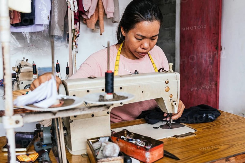 A asian seamstress focuses on sewing clothes in a workshop using a vintage sewing machine. Tools are scattered on a wooden table, and colorful fabrics hang in the background