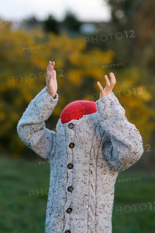 A young unrecognizable child, hiding while wearing a whimsical red hat and textured gray sweater, raises their hands playfully against an autumnal background in the park