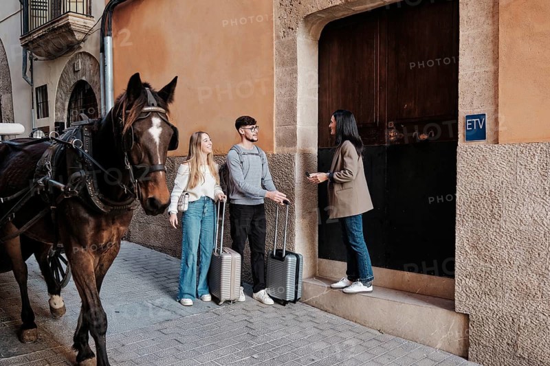 A young couple arrives with suitcases to explore their vacation home in Majorca, Spain. A woman welcomes them at the entrance while a horse stands nearby on the cobbled street