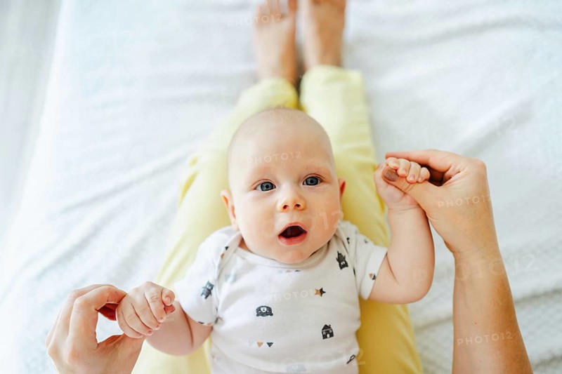 A baby in a patterned onesie lies on a mother lap, holding their hands. The setting is cozy and bright, capturing a moment of bonding and tenderness