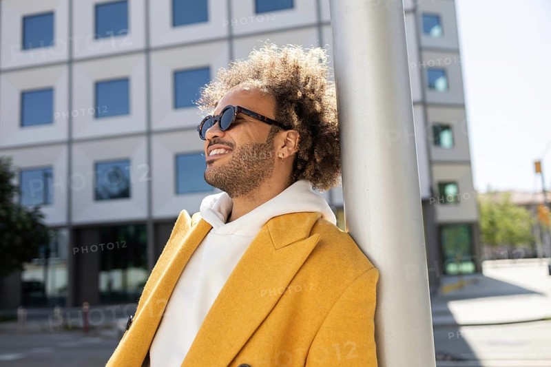 A confident Afro man in a bright yellow coat enjoying the sunshine, leaning against a pole in a cityscape. His relaxed demeanor captures a sense of freedom and leisure