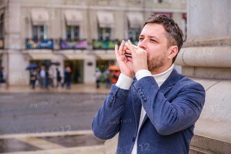 Man musician in a blue blazer and white sweater playing blues on a harmonica with eyes closed, standing outdoors in city street