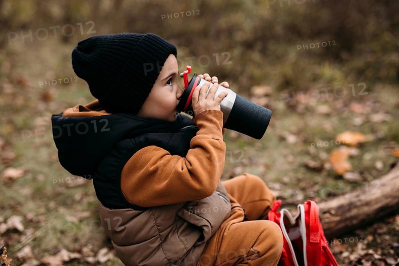 A child dressed warmly in a black beanie and brown outfit drinks from a thermos while sitting on the ground in an autumn park, surrounded by fallen leaves and a red backpack