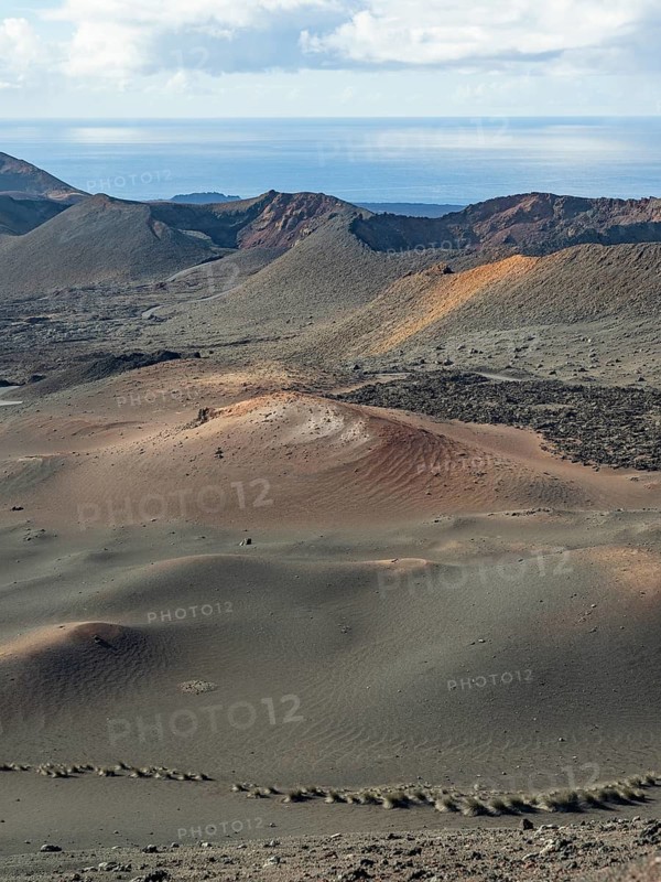 A stunning landscape in Timanfaya National Park, Lanzarote, showcasing volcanic terrain with rolling hills under a cloudy sky, set against the backdrop of the Atlantic Ocean