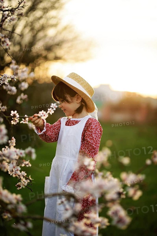A young girl wearing a white dress and straw hat admires almond blossoms at sunset in a lush garden, capturing the innocence and joy of spring in nature