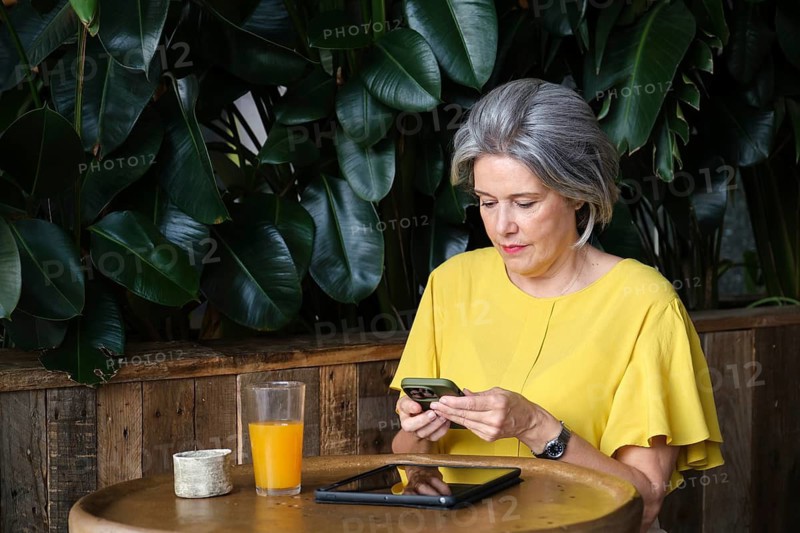 A mature woman with gray hair and a yellow shirt uses her smartphone while sitting at a wooden table in a cozy cafe, surrounded by lush green plants. A tablet and juice are on the table