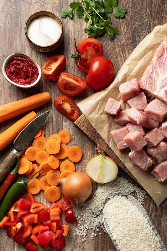 Top view of fresh ingredients arranged for preparing Hungarian pork goulash with rice. The image features raw pork cubes, rice, tomatoes, carrots, onion, sour cream, hot paprika paste, and herbs spread on a wooden surface